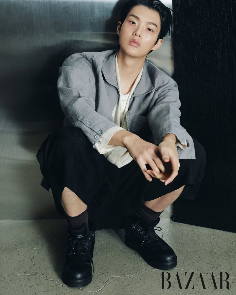 Model crouching against a metallic wall wearing a grey jacket, white top, black trousers, and dark boots.