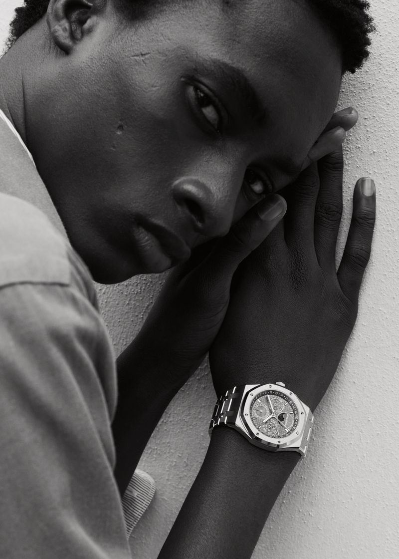 Close-up black and white portrait of a Black man leaning against a textured wall, wearing a luxury metal wristwatch.