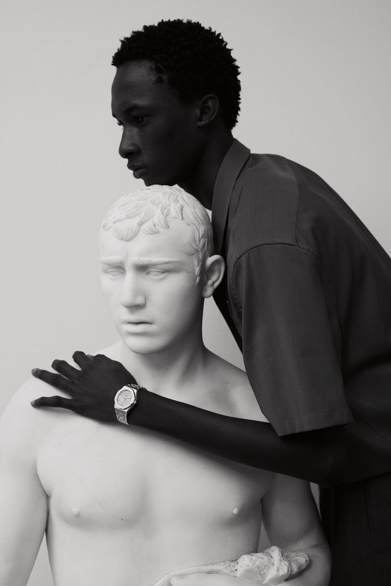 A Black man posing behind a white classical bust sculpture wearing a short sleeve shirt and silver wristwatch against a grey studio backdrop.