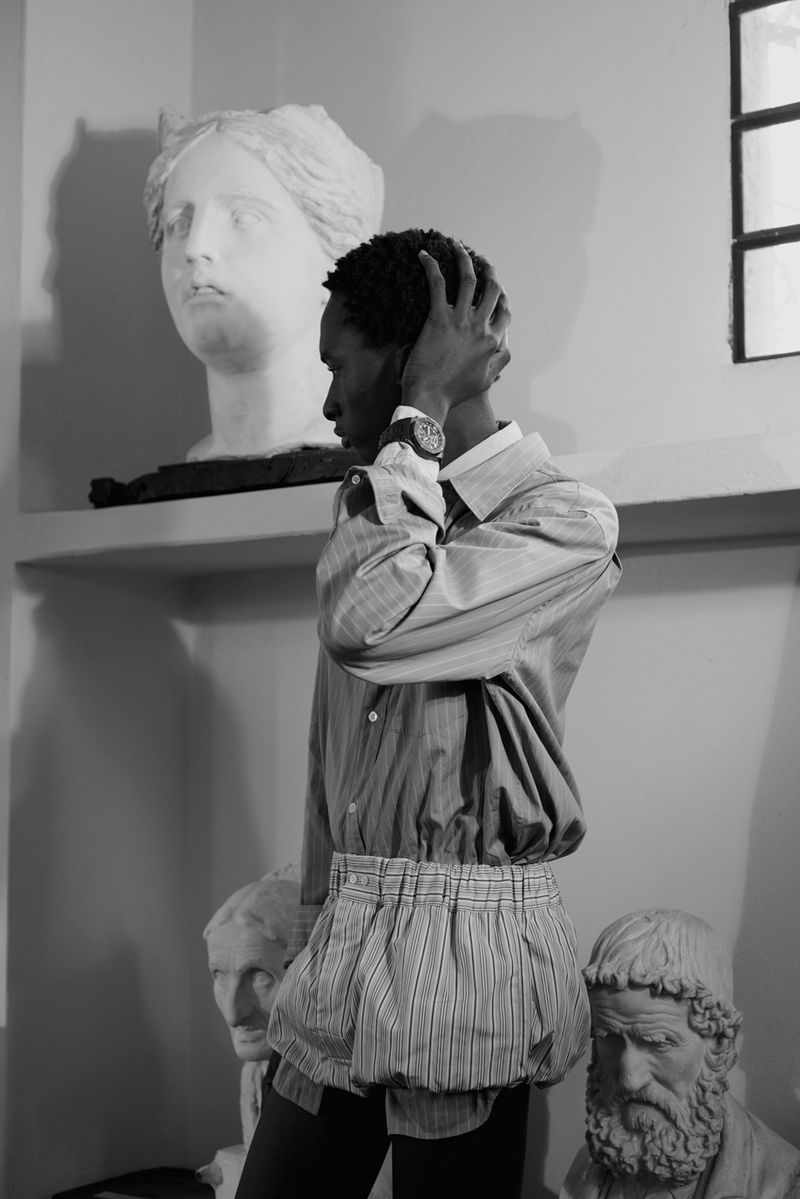 Black man wearing a striped shirt and wristwatch posing among classical marble sculptures in an editorial photograph.