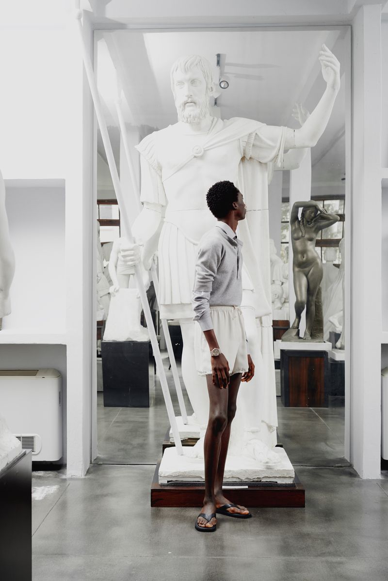 Black man standing in a museum gallery next to a large white classical statue holding a staff