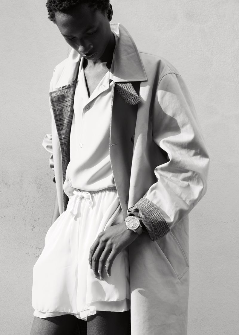 Black man wearing a trench coat and shorts with a luxury wristwatch against a plain wall.