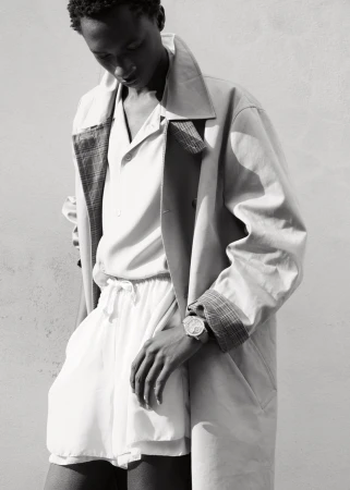 Black man wearing a trench coat and shorts with a luxury wristwatch against a plain wall.