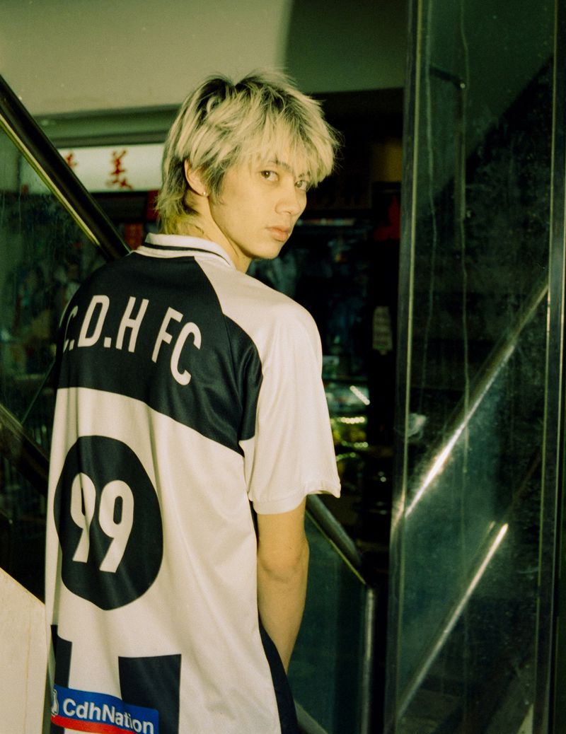 Young man with bleached blonde hair wearing a white and dark blue sports jersey looking back over his shoulder.