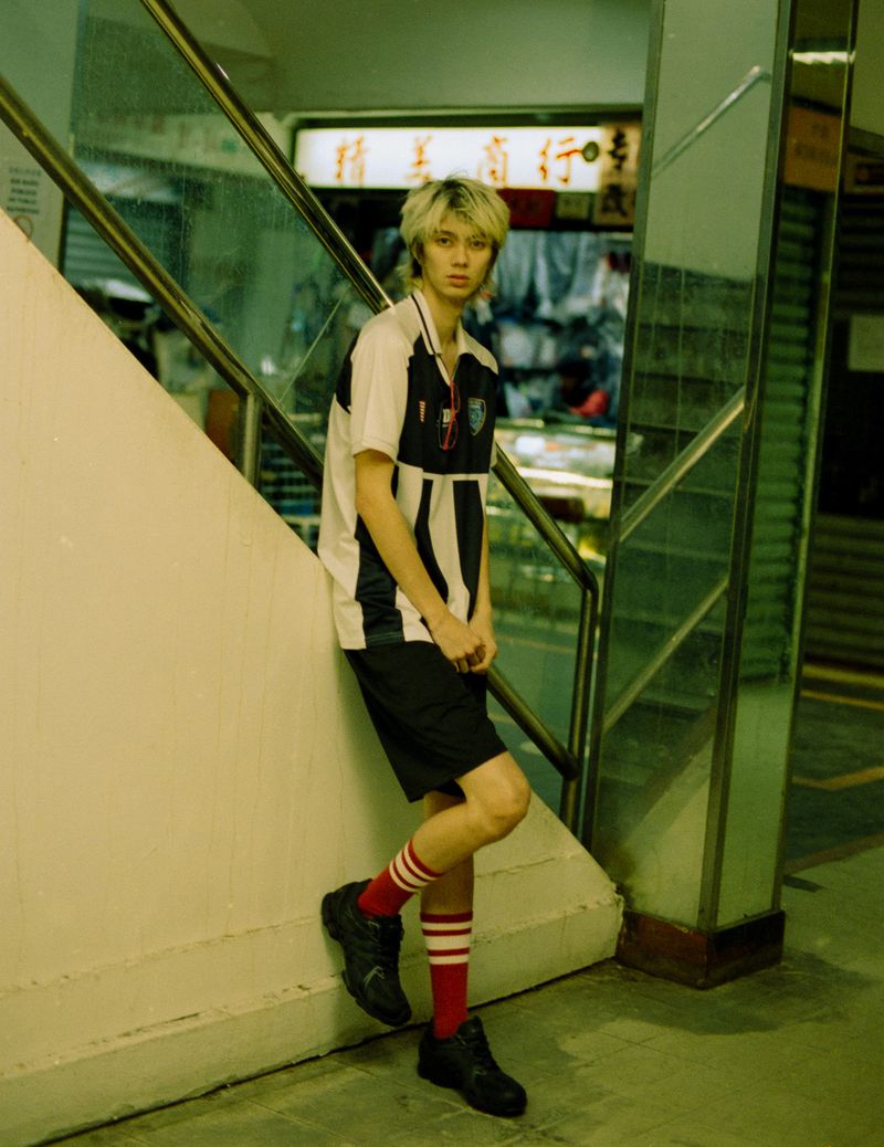 Young adult poses in black-and-white soccer jersey, black shorts, and red-and-white striped socks near a city stairway.