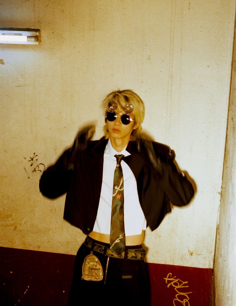 A blonde model wearing sunglasses, a black jacket, white shirt, and camo tie against a textured concrete wall.