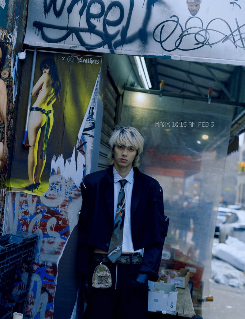 Young man with dyed blonde hair wearing a dark jacket and tie standing on a gritty city street