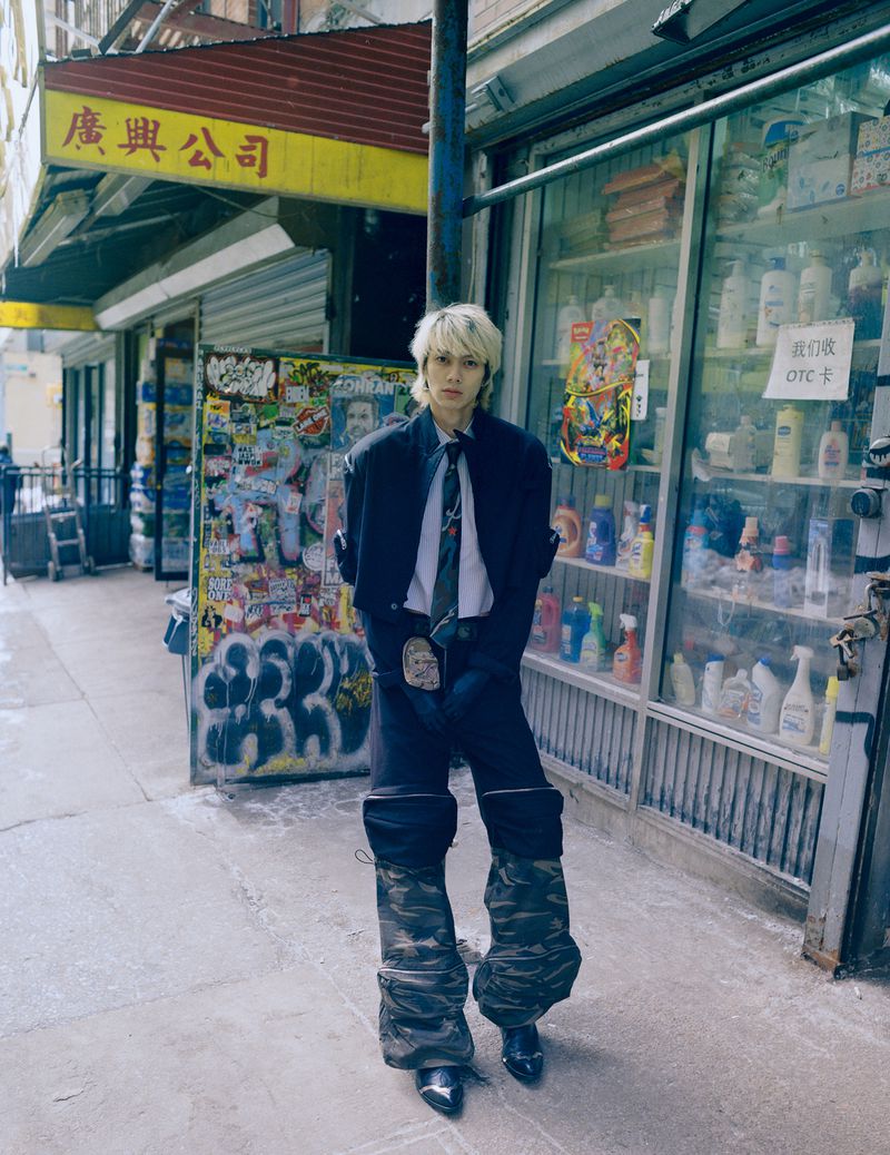 Model with blonde hair wearing dark jacket, white shirt, and camo trousers standing outside a storefront