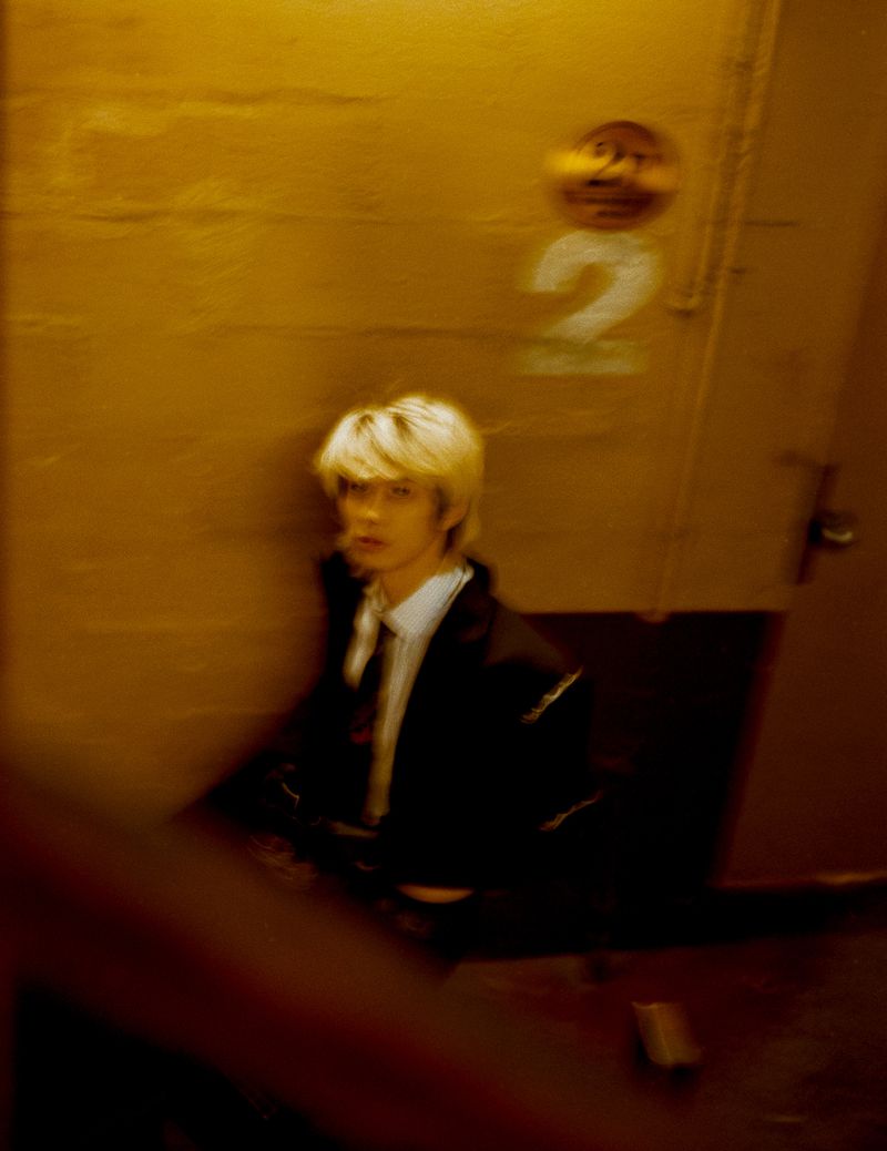 Blonde man wearing a dark jacket and tie in a dimly lit hallway with yellow walls and a number sign