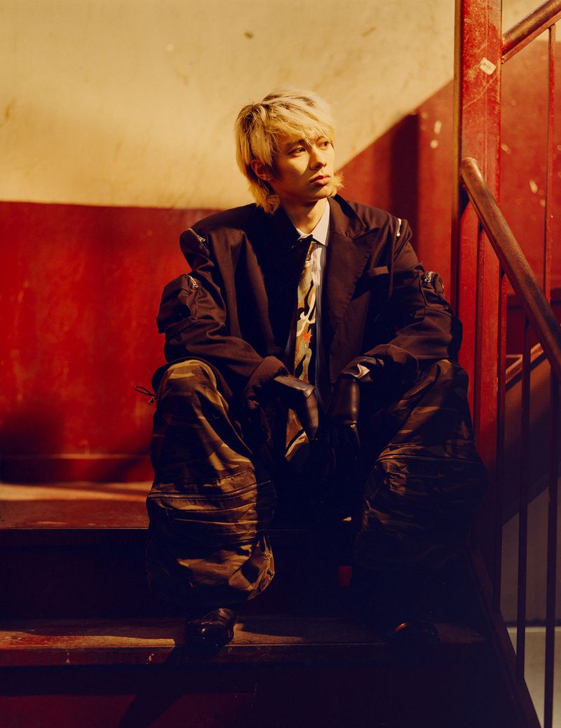 Blonde man wearing a dark blazer, patterned tie, and camo cargo pants seated on indoor stairs
