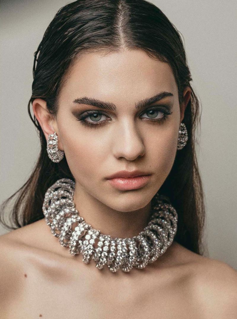 Close-up portrait of a woman wearing a heavy crystal choker necklace and matching earrings against a neutral grey studio backdrop.