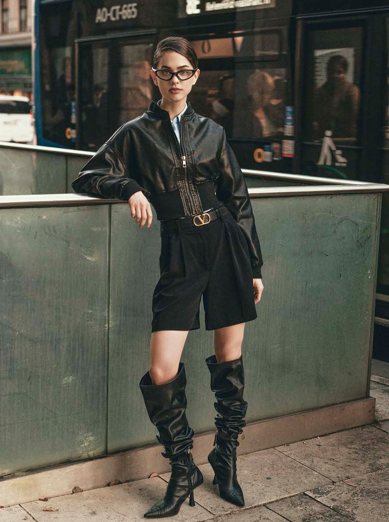 A woman in a black bomber jacket, high-waisted shorts, and black leather high-heeled boots, standing in an urban environment.