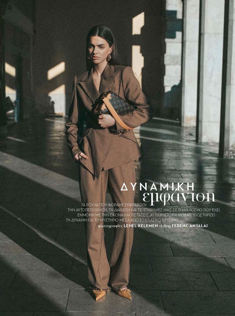 Model in a brown pantsuit holding a patterned handbag standing outdoors near stone columns in golden sunlight.