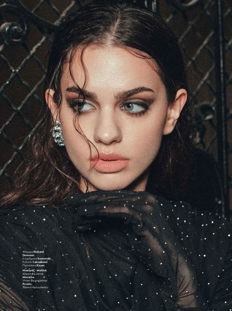 Close-up portrait of a woman with dark hair, sparkling sheer gloves, and rhinestone earrings against an ornate wrought iron background.