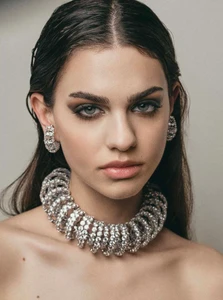 Close-up portrait of a woman wearing a heavy crystal choker necklace and matching earrings against a neutral grey studio backdrop.