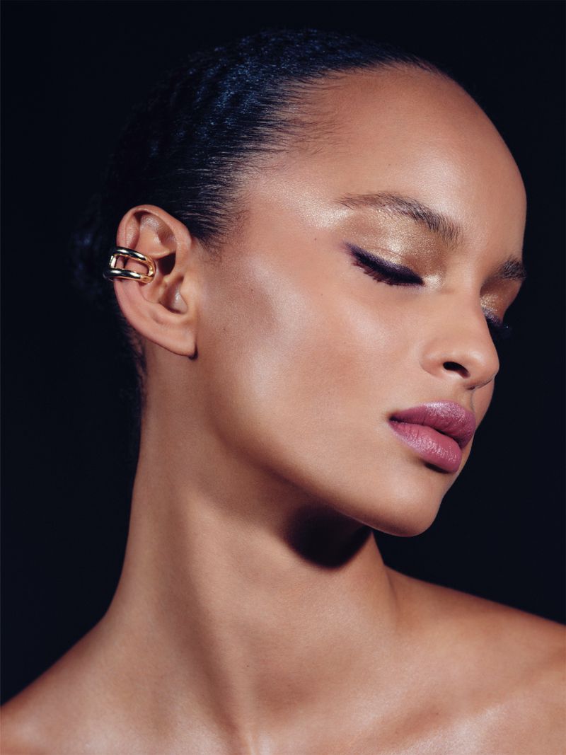 Close-up portrait of a woman wearing gold ear cuffs and makeup against a black background for Hermes Beauty in Elle UK.