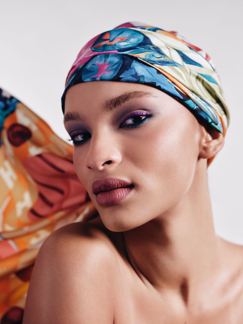 Close-up portrait of a woman wearing a colourful patterned headscarf and vibrant purple eyeshadow against a neutral background
