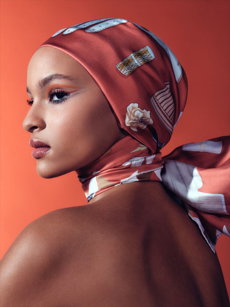Close-up studio shot of a woman wearing a patterned headscarf, featuring warm orange and white tones against an orange backdrop.