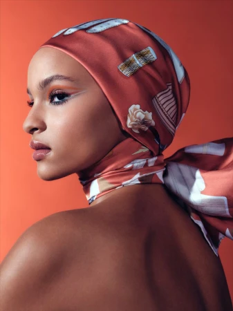 Close-up studio shot of a woman wearing a patterned headscarf, featuring warm orange and white tones against an orange backdrop.