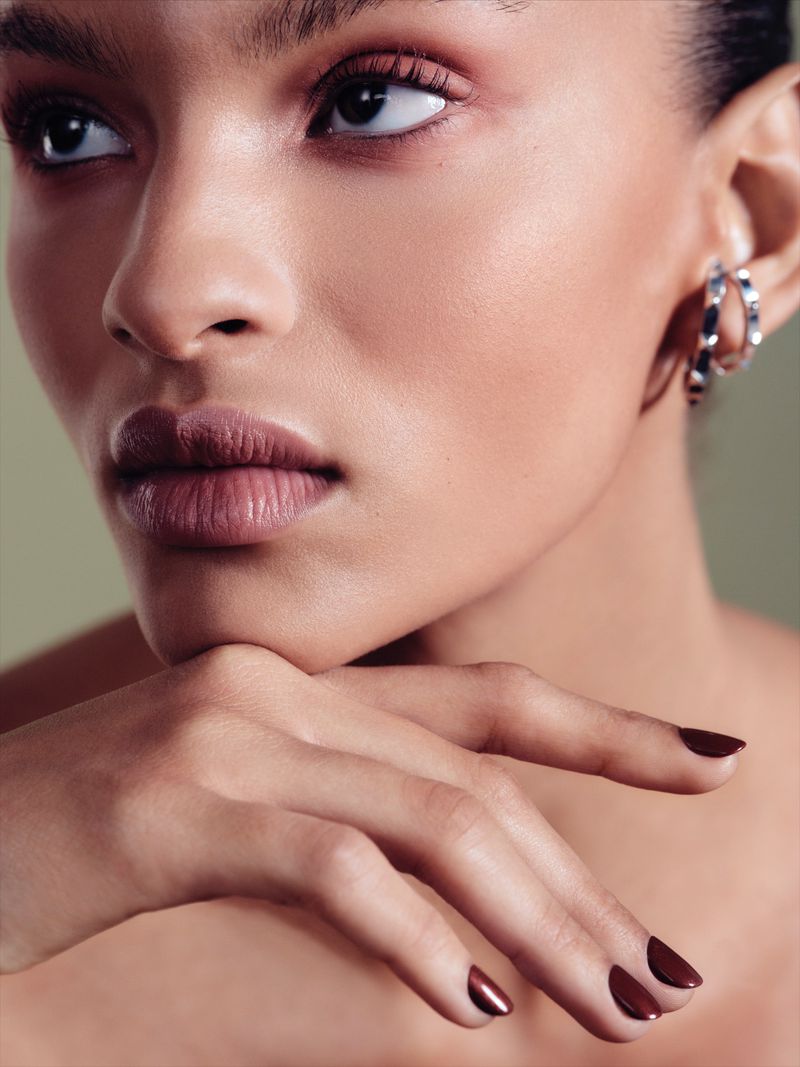 Close-up portrait of a model resting her chin on her hand with dark nail polish, wearing silver ear cuffs.