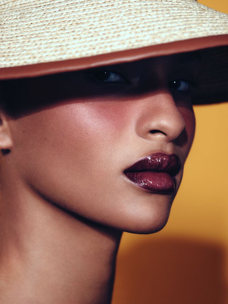Close-up portrait of a model wearing a wide-brimmed straw hat and glossy dark lipstick against a warm background.