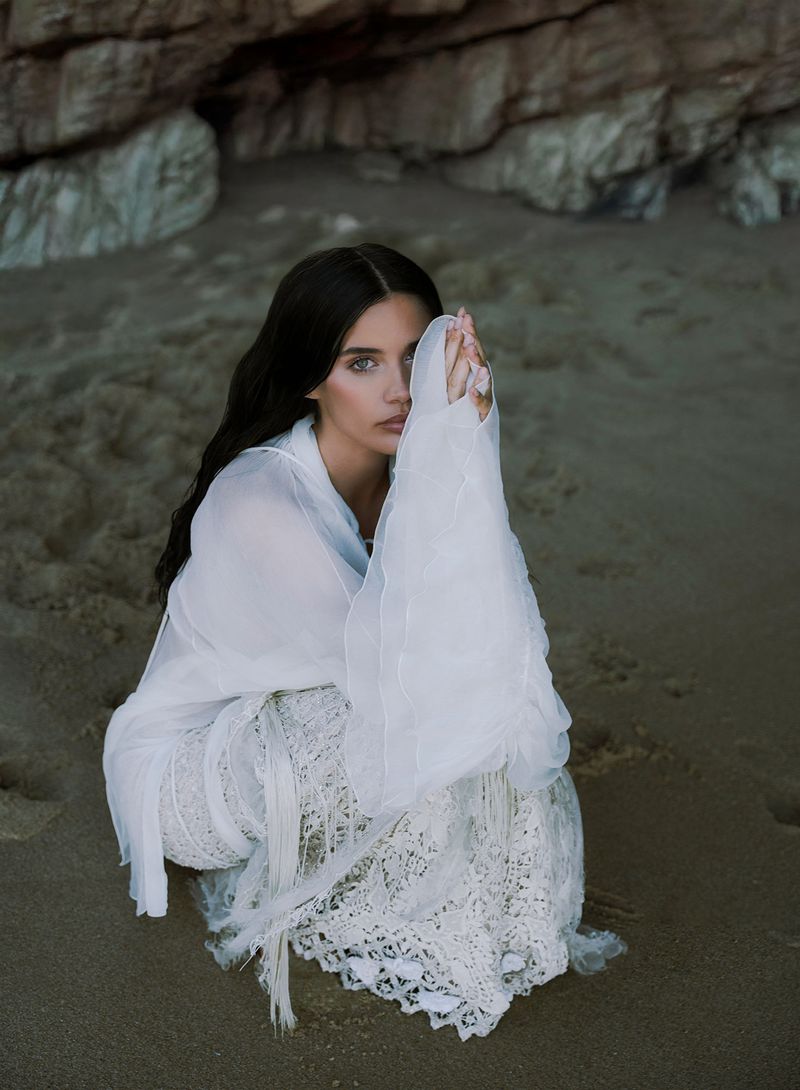 Woman in white lace dress sits on a sandy beach with rocks in the background, covering part of her face with her hands.