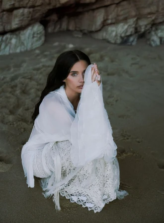 Woman in white lace dress sits on a sandy beach with rocks in the background, covering part of her face with her hands.