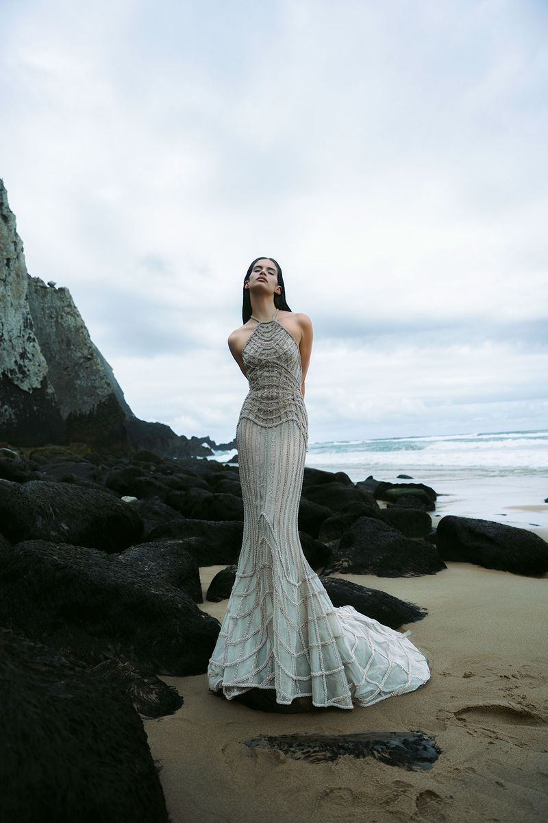 Woman in gown on sandy beach with rock formations. Long beaded halterneck dress with train, eyes closed, sky is overcast.