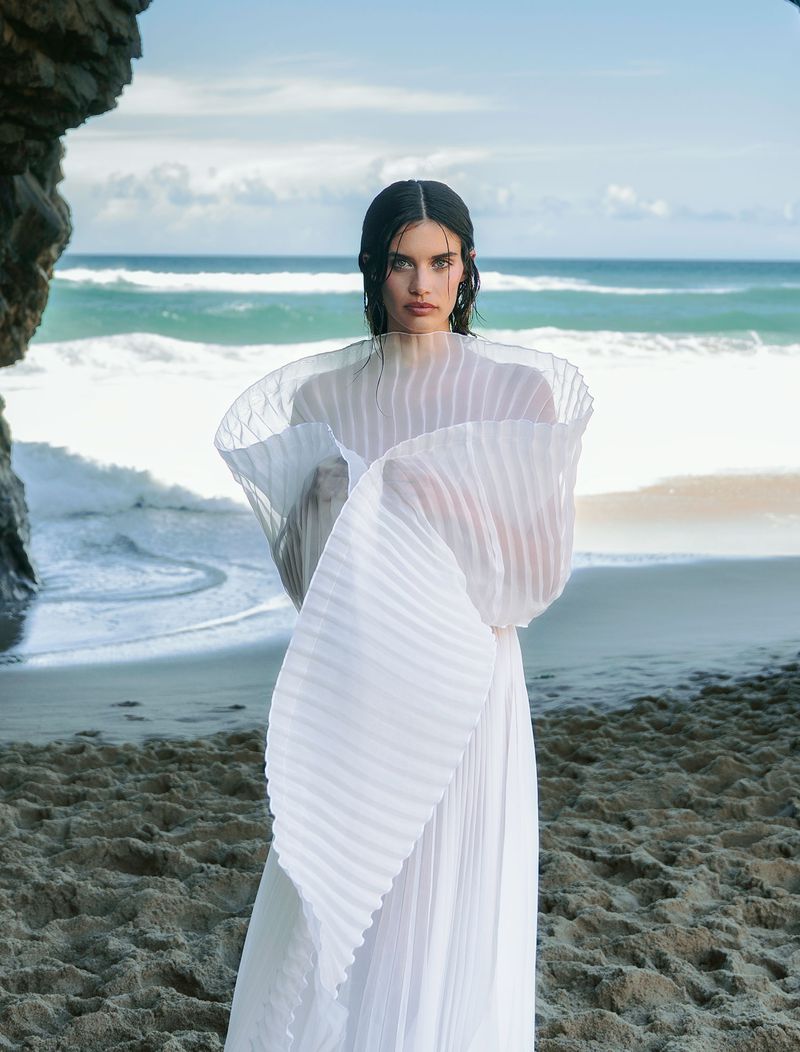 A woman in a flowing white dress stands on a sandy beach with crashing waves and a rocky outcrop in the background, creating a serene fashion scene.