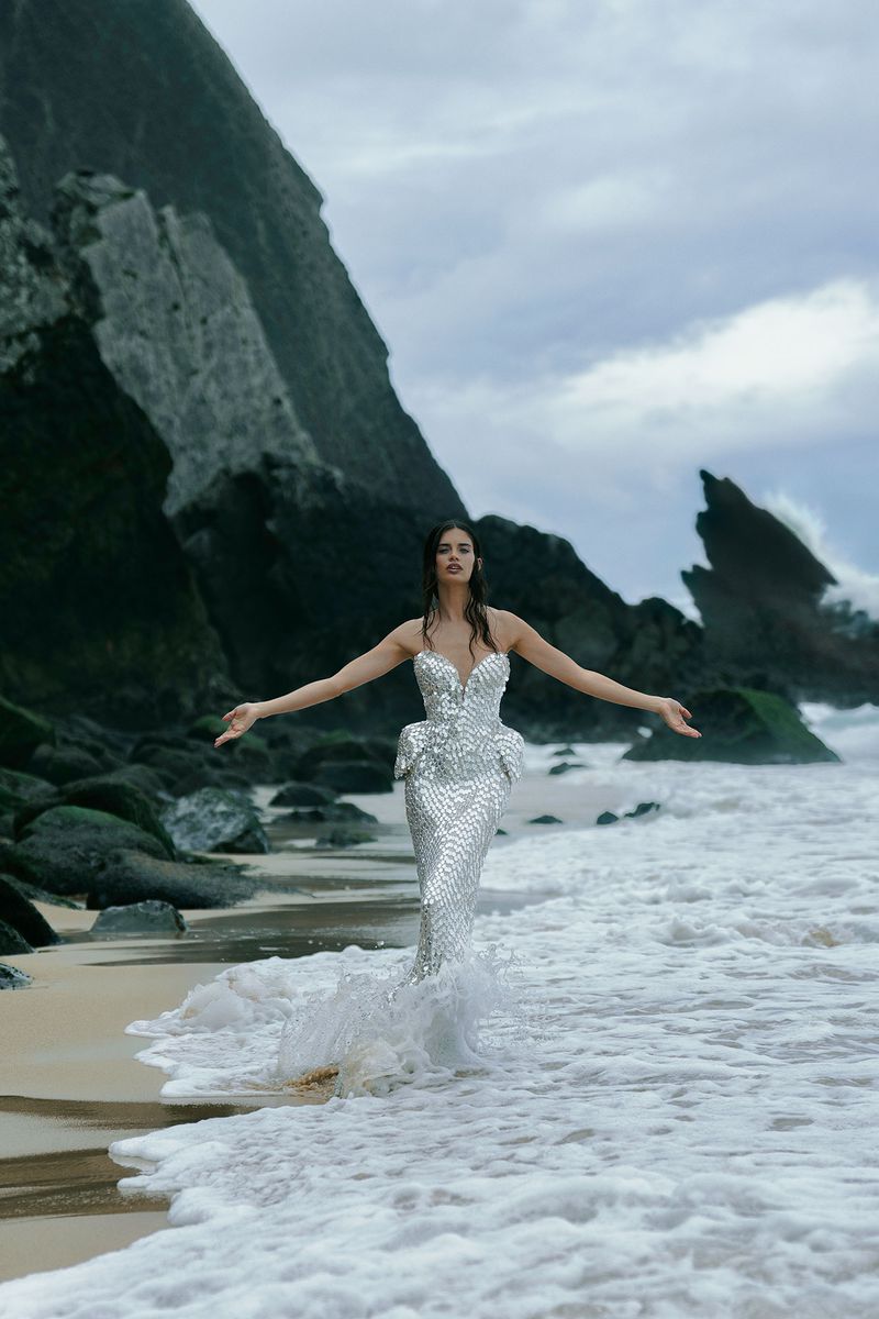 Woman in silver gown stands in surf on beach with rocky cliffs and sky. Ocean waves with light reflection, arms open. Fashion shoot.