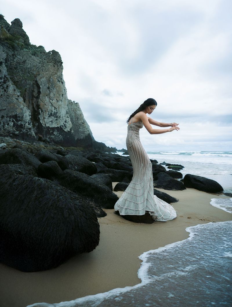 Woman in gown stands on a beach near rock formations; ocean waves roll in under an overcast sky, in a full shot.