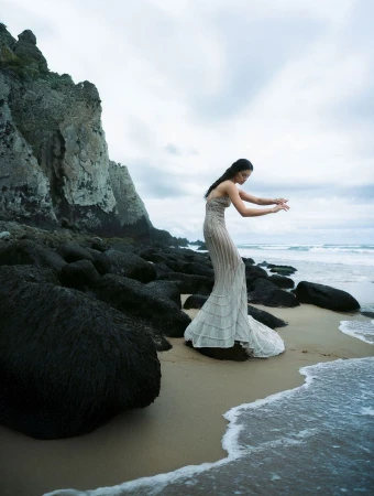 Model Sara Sampaio posing in a strapless textured grey knit gown on a rocky beach by the ocean for Vogue Portugal.