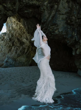 Model Sara Sampaio in a flowing white sheer top and fringed crochet skirt standing on a rocky beach with arms raised.