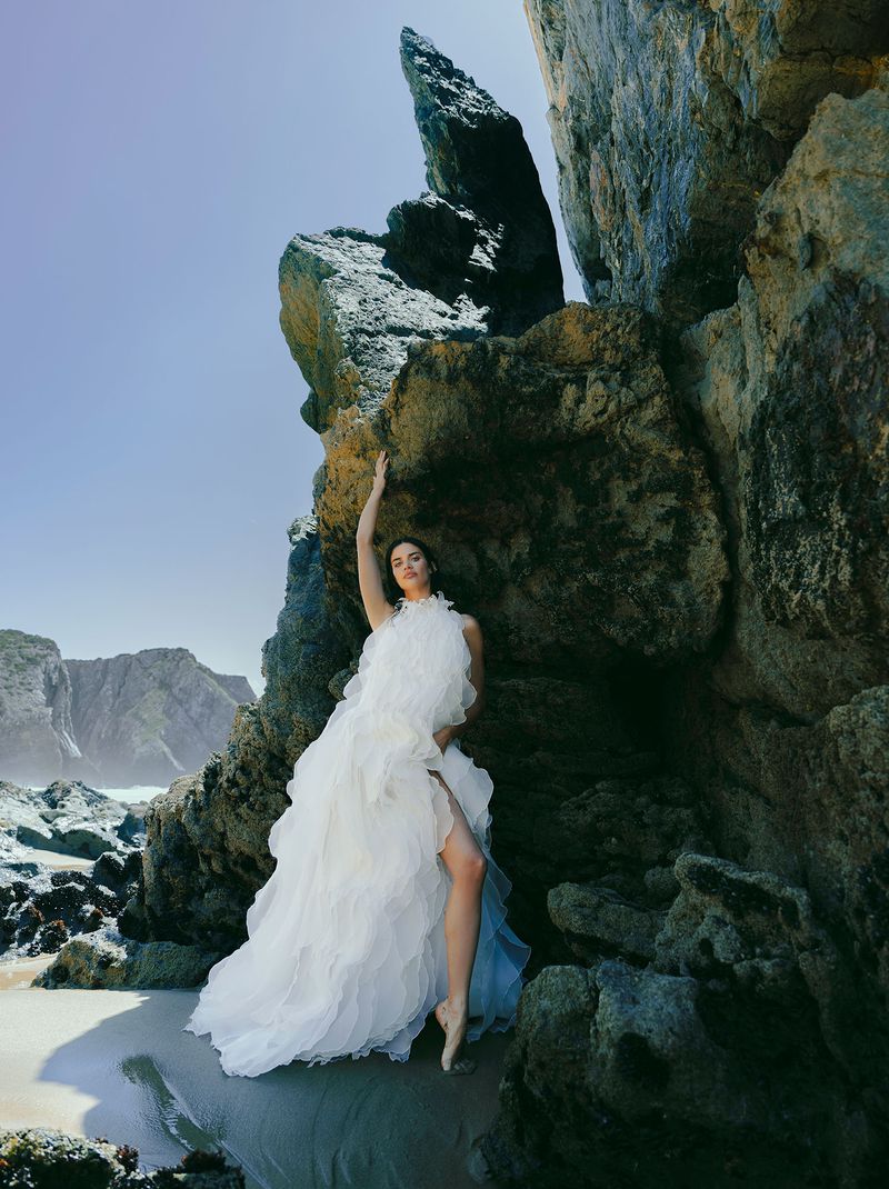 Woman in a white ruffled gown poses barefoot on a beach near rock formations under a bright sky, creating a striking contrast.