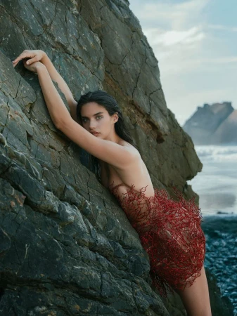 Woman in a decorative red dress leans against a large rock on a beach with ocean and distant mountains, posing for a fashion shot.