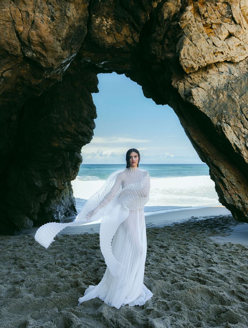 A woman in a white dress stands on a beach framed by a rock archway with ocean and sky visible in the background, bathed in natural light.