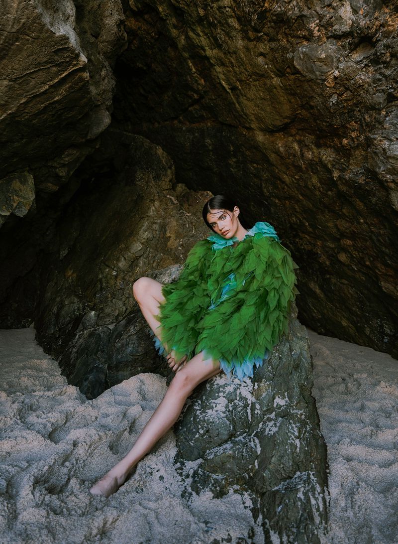Woman in green feathered coat sitting on a rock in a cave with sandy ground. Fashion editorial shot with natural lighting, outdoor setting.