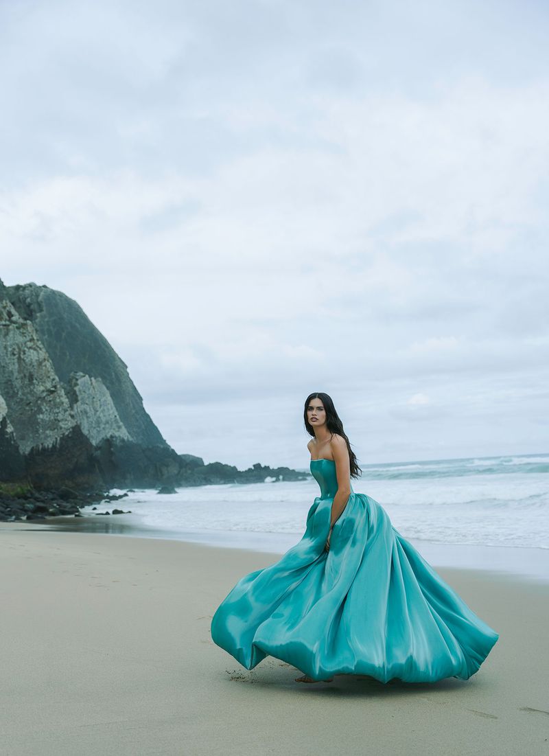 Woman in a teal ballgown standing on a sandy beach with the ocean behind her and cliffs in the distance under a cloudy sky.