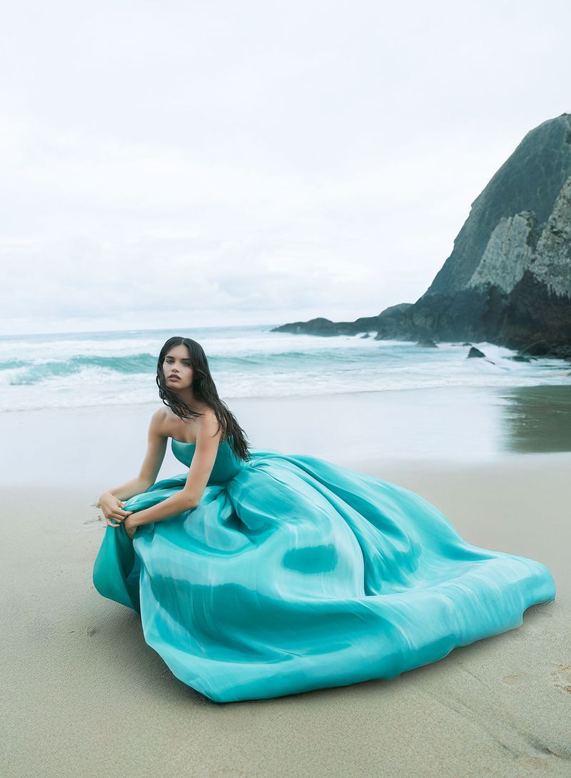 A woman in a blue strapless gown sits on a sandy beach with ocean waves and a rocky coastline under an overcast sky.