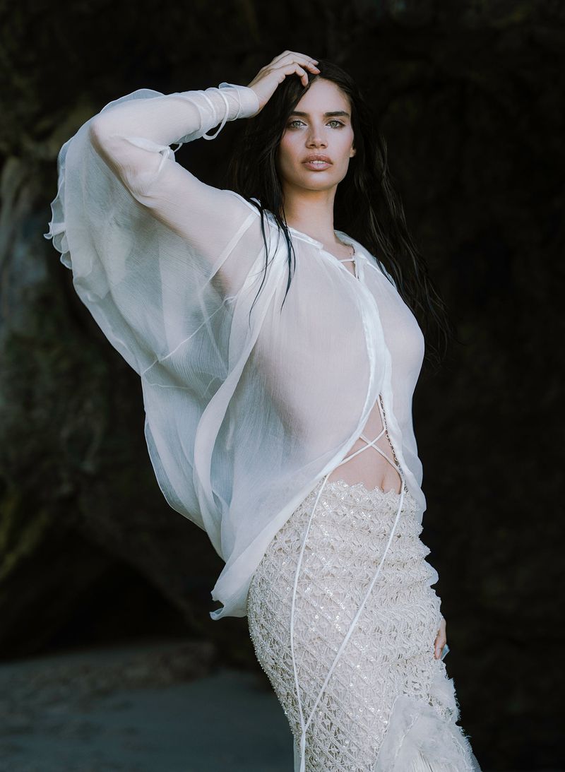A woman with long dark hair poses in a sheer white top and embellished white skirt against a dark rocky backdrop in a fashion shot.