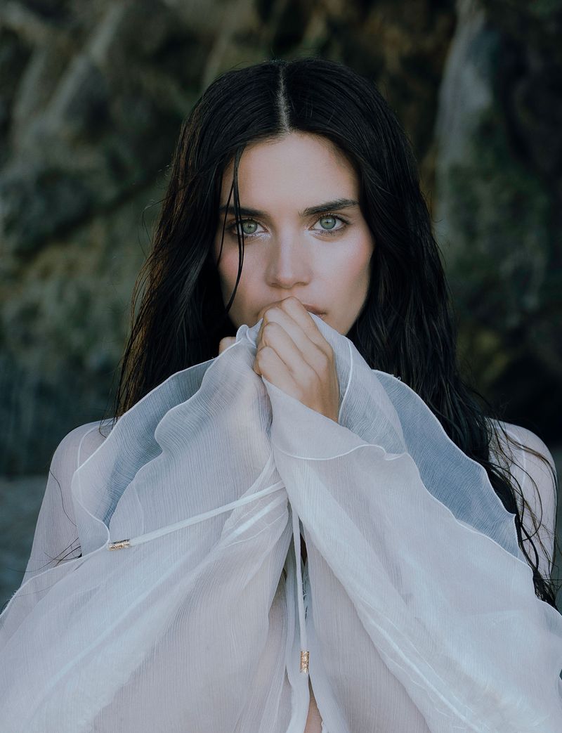 Striking portrait of a young adult woman with dark hair and light eyes, holding a sheer white fabric against a dark, rocky background.