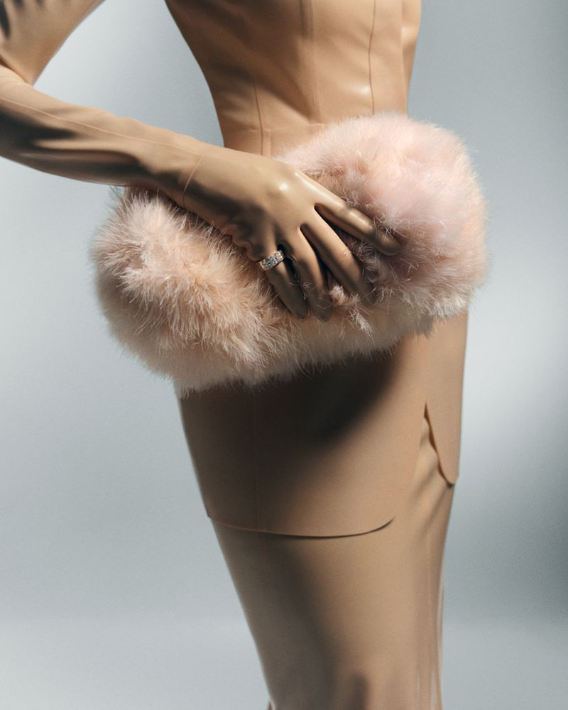 Close-up of a person wearing a tan leather dress and holding a fluffy pink faux-fur clutch against a light grey background.