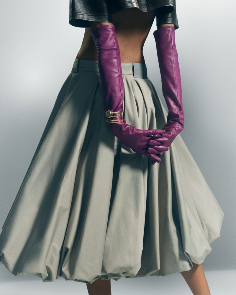 Model wearing a pleated grey midi skirt, black leather top, and long purple leather opera gloves for L'Officiel USA