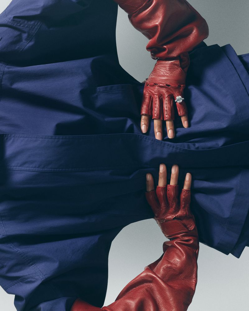 Close-up of hands wearing red leather gloves and a diamond ring against a blue garment on a light grey background.