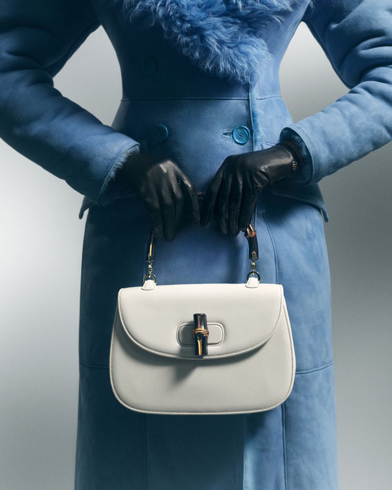 Close-up of a person wearing a blue coat and black gloves holding a white handbag with a bamboo handle