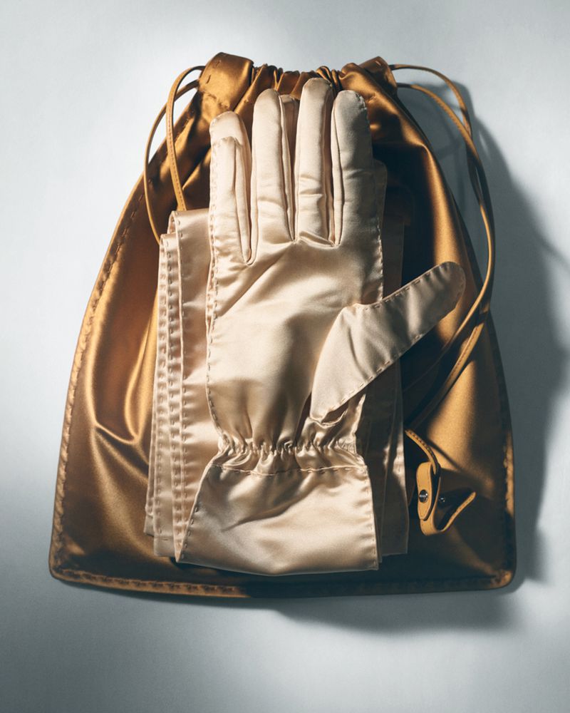 Close-up product shot of shiny satin gloves resting on top of a matching gold drawstring dust bag against a white seamless background.
