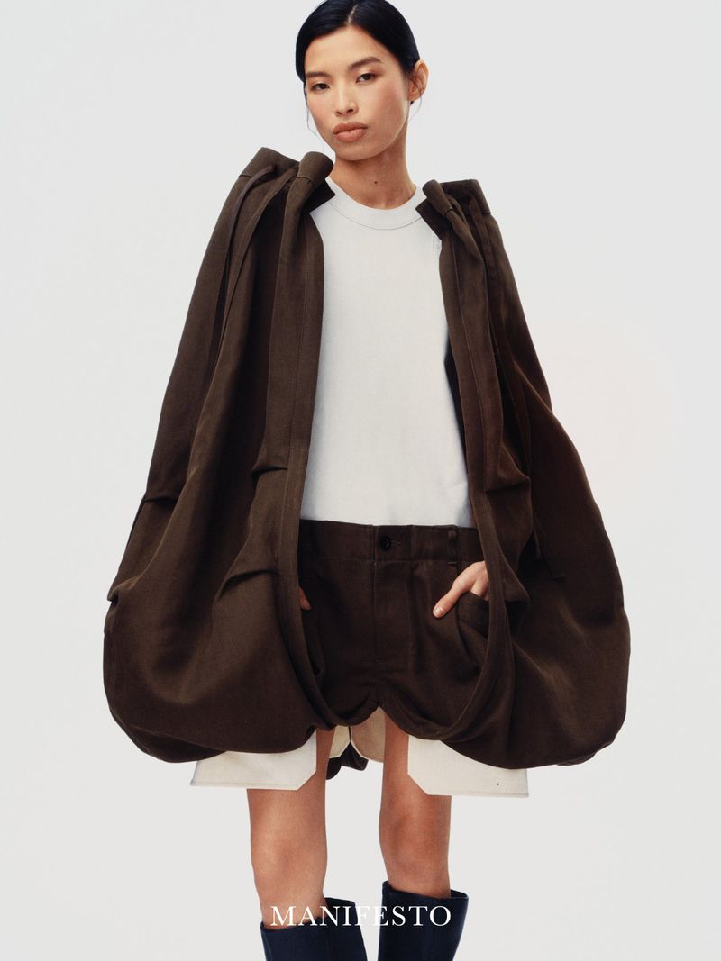 Model wearing a voluminous dark brown coat and white t-shirt against a plain white background for Manifesto Magazine