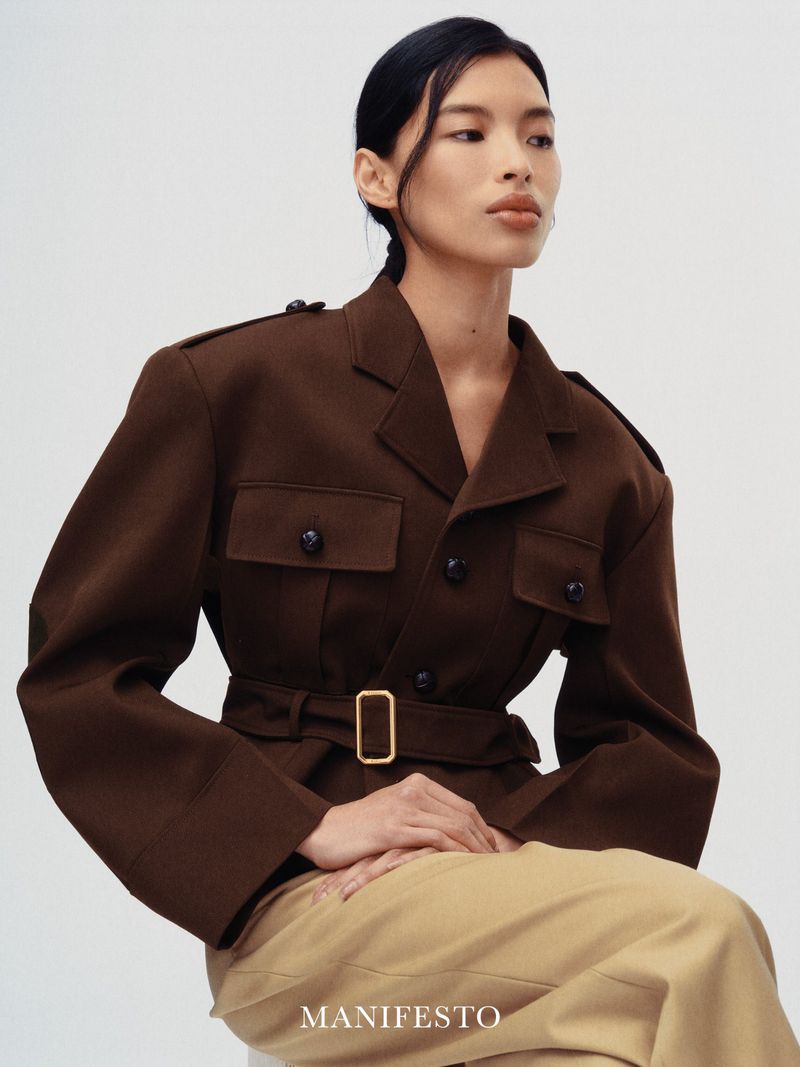Model wearing a dark brown structured belted jacket and tan trousers against a plain white background for Manifesto Magazine.