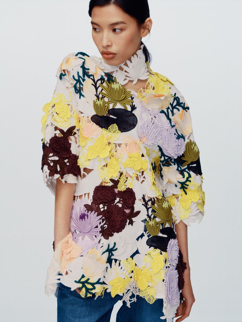 Model wearing a white sheer tunic covered in colorful floral appliqués and blue jeans against a neutral background.