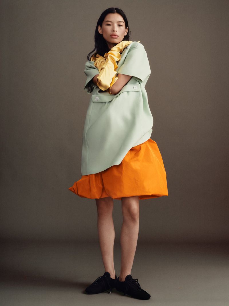 Model wearing a pale green top with orange skirt and black sneakers against a neutral grey studio background